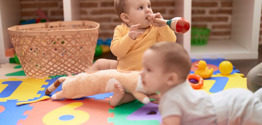 Two toddlers sucking maraca sitting on floor at kindergarten